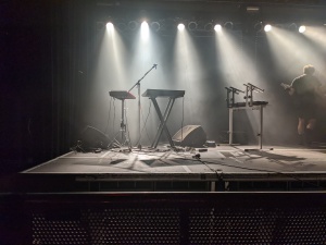 Bühne im Umbaulicht. 2 Synthesizer stehen da, mit Mikrofonständer. Rechts daneben steht noch der Aufbau eines anderes Synthesizers, der gerade hinter die Bühne getragen wird von einem Helfer.
