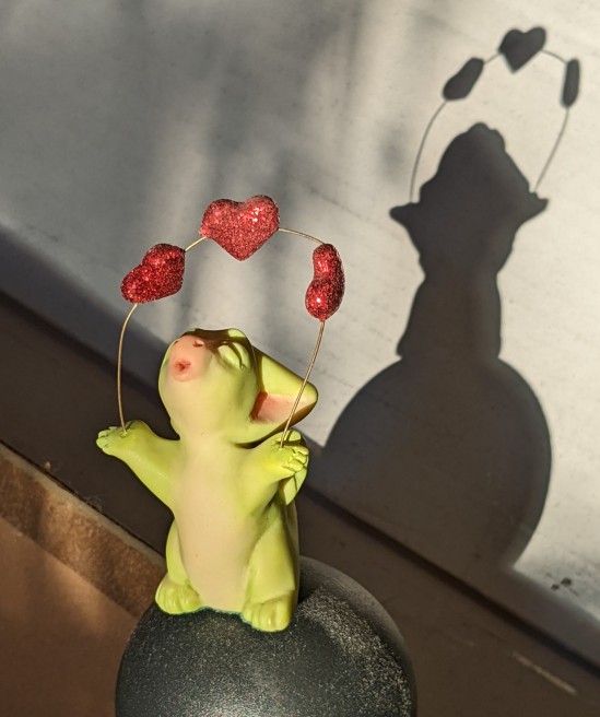 Eine kleine grüne Drachenfigur hat drei rote Herzchen über sich und wirft einen Kussmund in die Luft - im Hintergrund ist der Schatten der Figur zu sehen
