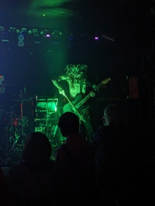 Im grünen Bühnenlicht steht ein Mann mit nacktem Oberkörper und Maske mit Gitarre auf der Bühne, die er mit einem Bogen streicht